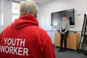 A youth worker in a red hoodie with 'Youth Worker' on the back watches a young person present to the group