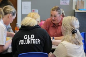 A girl sits at a table wearing a black hoodie with 'CLD Volunteer' on the back