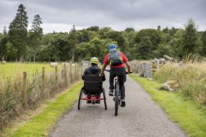 Two cyclists, one in an adapted bike, cycle on a country lane away from the camera