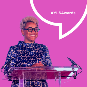 Finalists announced for YLS Awards 2024. A previous presenter stands at a podium speaking and smiling with a pink background and speech bubble behind them.