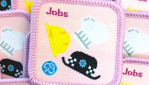 Stitched pink badge with the word 'Jobs' in thetop left and three hats - construction worker, chef and police, and a Girlguiding logo on it.