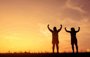 Outline of two children with their arms raised against a setting sun