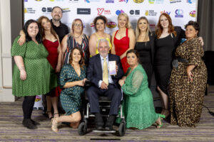 Group shot of awards winners at the national youth work awards. One man in a wheelchair at the front of the photo holds the trophy.