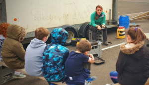Picture of a woman speaking to a group of young people sitting in front of a trailer.