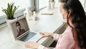 Woman with face mask talks to someone over a laptop, also wearing a facemask